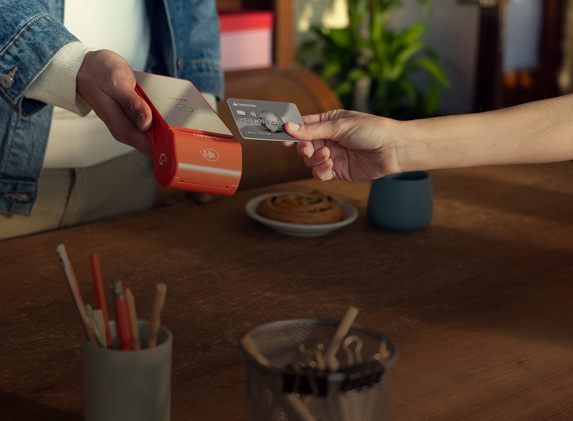 Mano acercando tarjeta para pagar en la terminal roja de Getnet que sostiene otra persona, sobre mesa con lápices y taza.