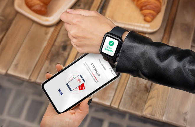 Mano sosteniendo un teléfono móvil con pantalla que muestra pago aprobado con tarjeta Visa, junto a un smartwatch con mensaje de confirmación de pago sobre una mesa con croissants.