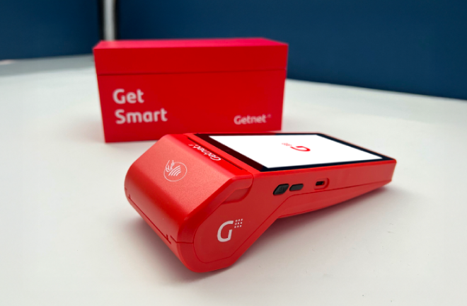 Terminal de cobro Getnet roja apoyada sobre superficie blanca, con pantalla encendida mostrando el logo y caja roja detrás con texto 'Get Smart'.