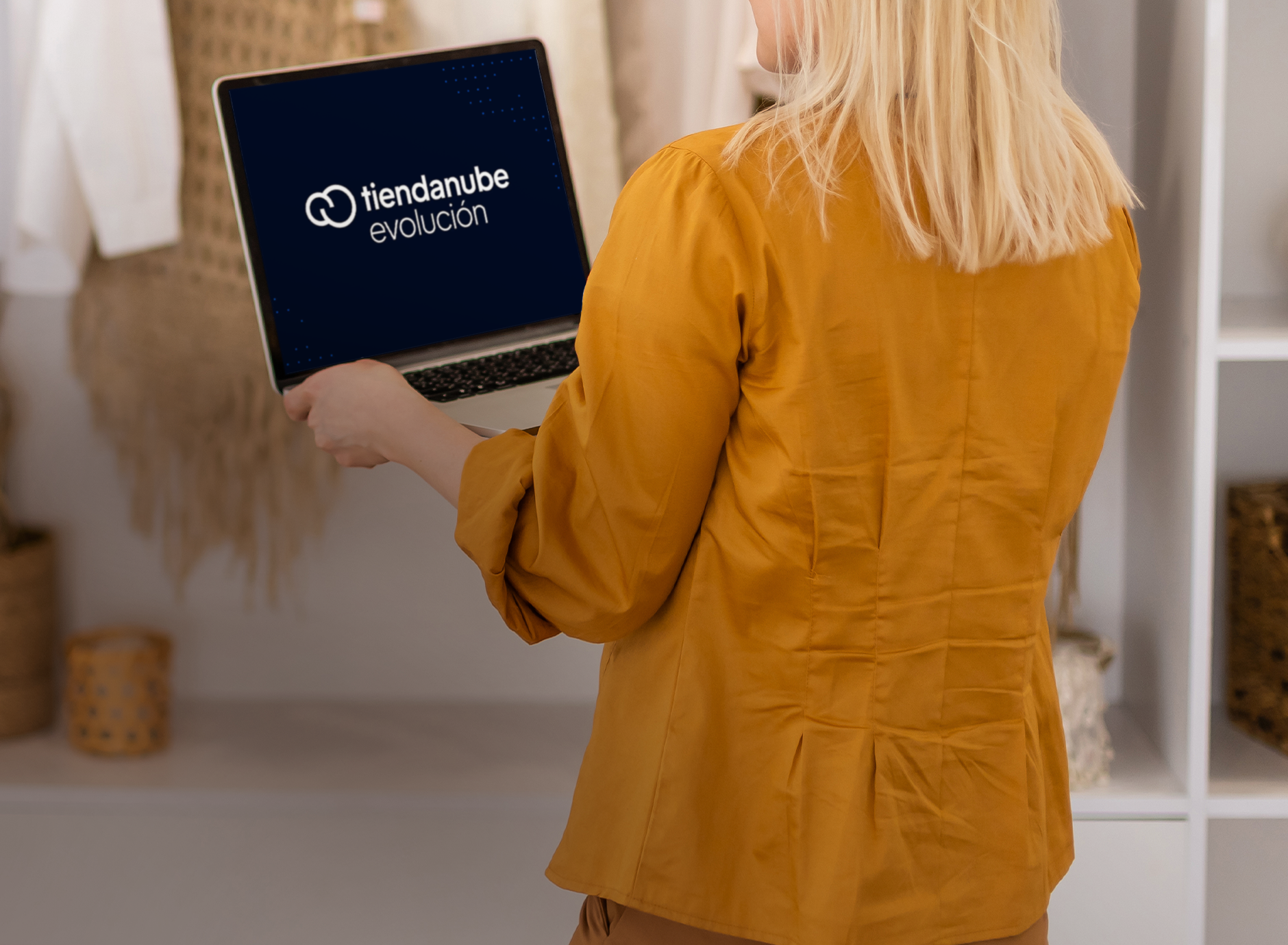 Persona sosteniendo una laptop que muestra el logo de Tiendanube Evolución, en un entorno con prendas colgadas.