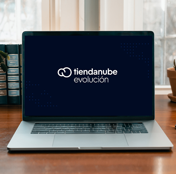 Laptop sobre un escritorio mostrando en la pantalla el logo de Tiendanube Evolución sobre fondo oscuro, con libros detrás y una maceta al costado.