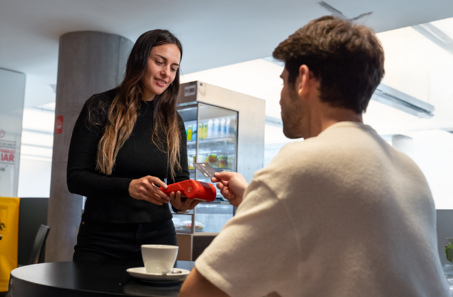 Persona acercando una tarjeta para pagar en una terminal de cobro Getnet roja en una cafetería, con taza sobre la mesa.