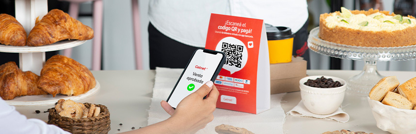Persona mostrando en un smartphone la confirmación de venta aprobada, frente a un cartel rojo de Getnet con código QR y mensaje 'Escaneá el código QR y pagá', sobre una mesa con croissants, galletas y una torta