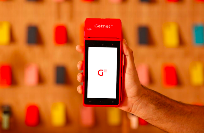 Mano sosteniendo una terminal de cobro Getnet roja con pantalla encendida mostrando el logo, sobre fondo con figuras coloridas.