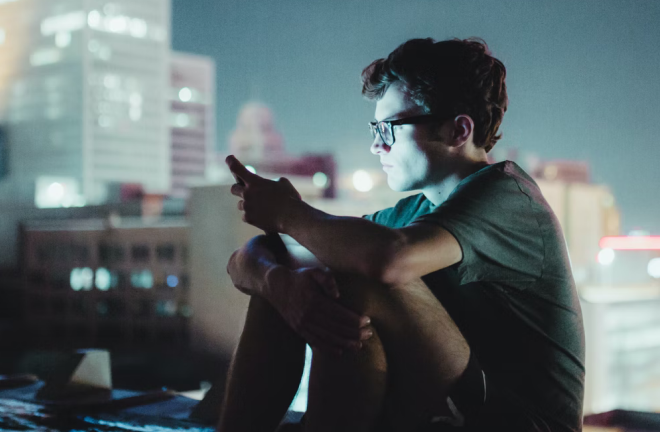 Persona sentada por la noche, sosteniendo un teléfono móvil, con luces y edificios desenfocados en el fondo.