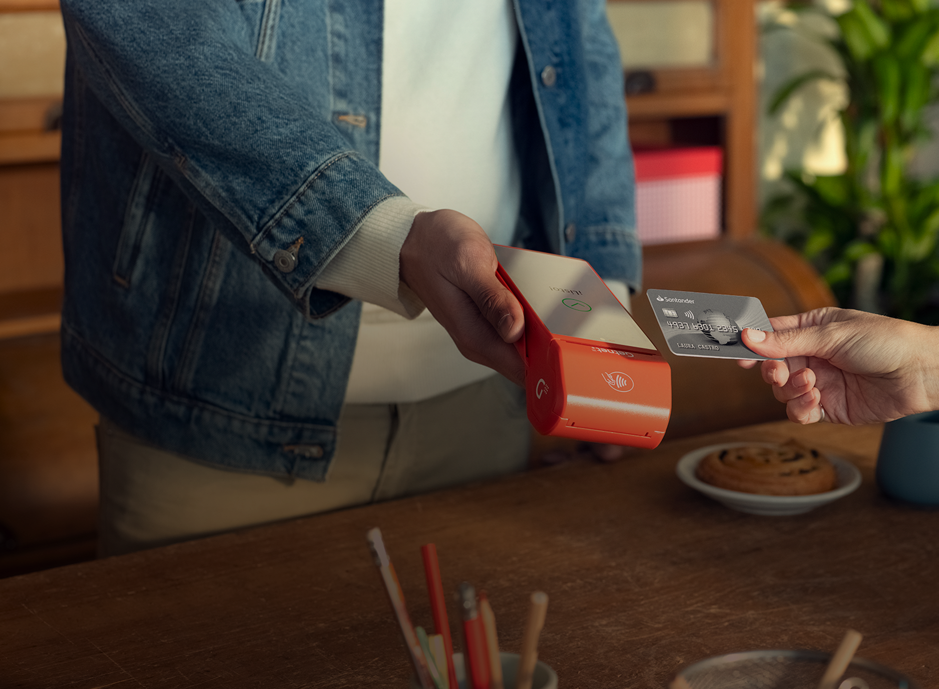 Mano acercando tarjeta para pagar en la terminal roja de Getnet que sostiene otra persona, sobre mesa con lápices y taza.