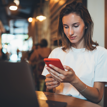 Persona usando un celular mientras trabaja con una laptop en una mesa de un café o restaurante.