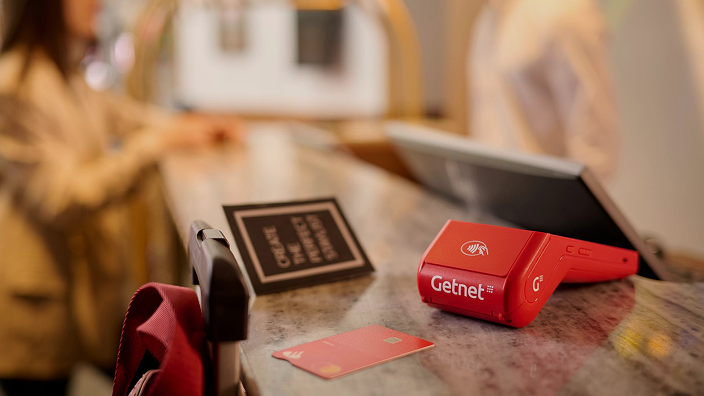 Terminal roja Get Smart POS de Getnet sobre el mostrador de un hotel, con una tarjeta y personal de recepción al fondo.