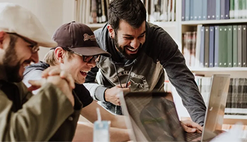 Tres hombres sonriendo por algo que ven en un portátil en una biblioteca