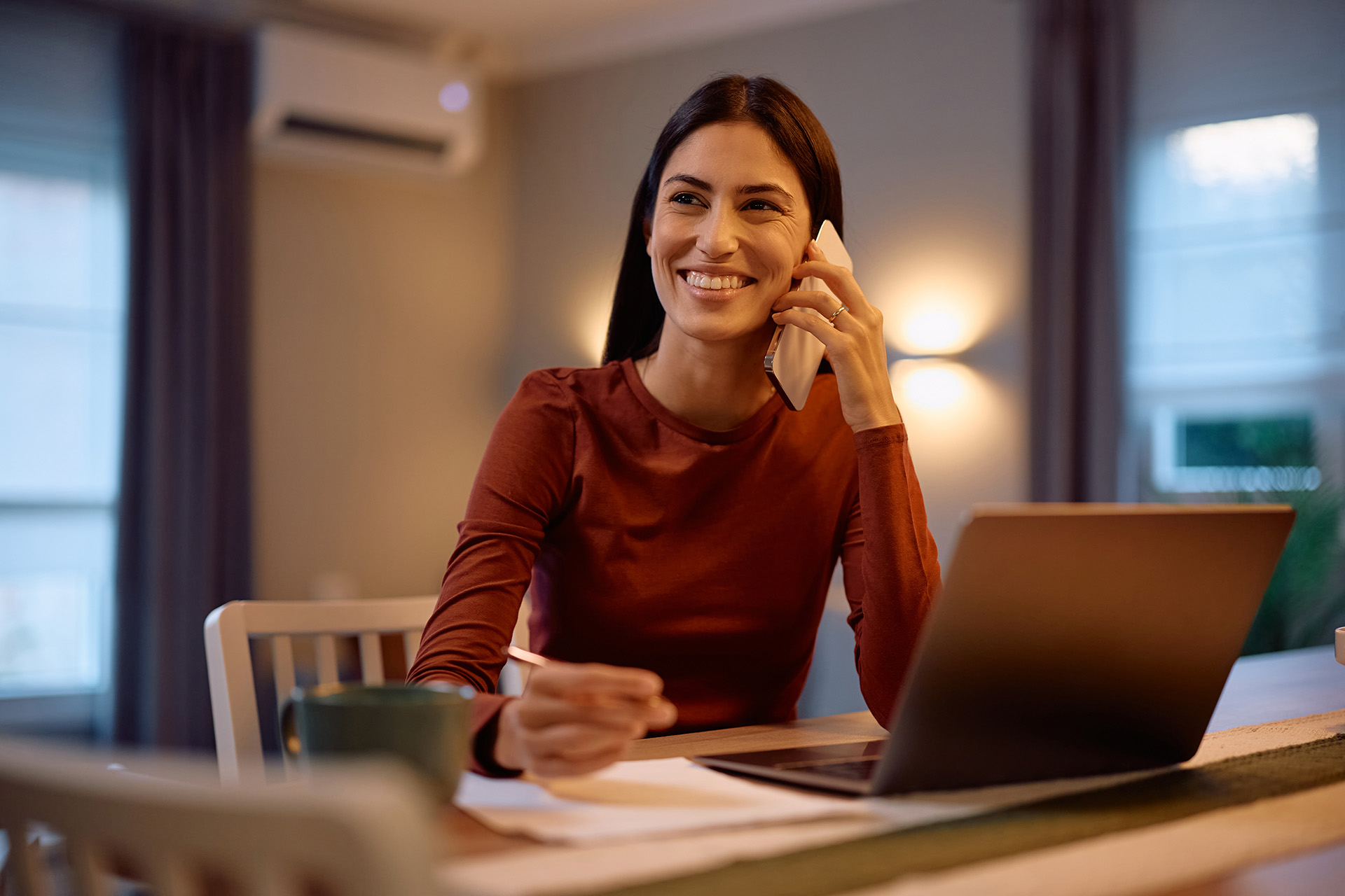 Mujer sonriendo con portátil en mesa, con una mano escribiendo y con la otra al teléfono 